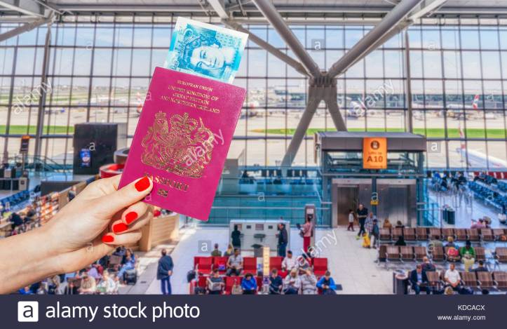 hand-holding-uk-passport-and-uk-5-pound-note-with-busy-airport-waiting-KDCACX
