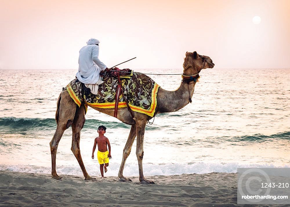 Boy stares in amazement at Arabic man wearing a thawb riding a camel on a beach, Dubai, UAE