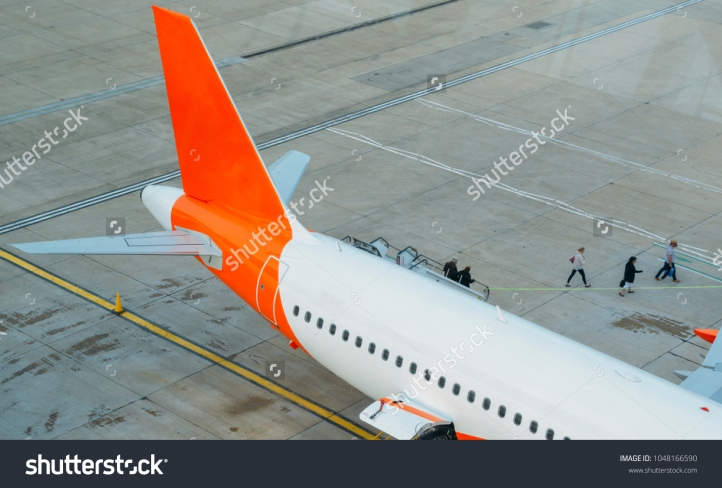 stock-photo-passengers-disembark-from-an-airplane-and-walk-on-tarmac-towards-the-terminal-building-1048166590